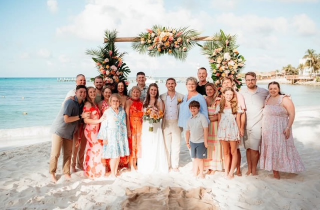 Wedding party and family gathered under a floral arch on a Caribbean beach.