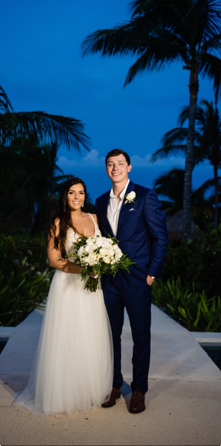 Couple in a white gown and navy suit standing beneath palm trees at blue hour.